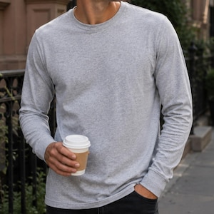 May include: A light grey long-sleeve crew neck shirt made of a heathered fabric. The shirt features a simple, solid-coloured design with long sleeves and a rounded neckline, providing a clean and straightforward appearance for casual wear.