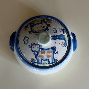 May include: A round ceramic serving dish with a lid. The dish is white with a blue rim and handles. The lid features a hand-painted design of a cow and other whimsical elements in blue and brown.