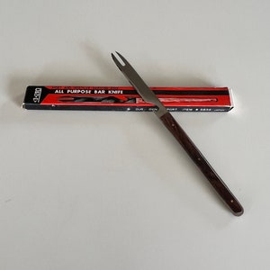 Op de afbeelding: Een zilveren barknife met een houten handvat. Het mes ligt op een wit oppervlak en heeft een rood-zwarte doos met de tekst "MOST AMNESTIC SERVING TOOL ALL PURPOSE BAR KNIFE" erop.