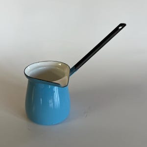 May include: A light blue enamel coffee pot with a black handle. The pot has a white interior and a rounded base. The handle is long and straight, with a small hole at the end. The pot is designed for stovetop use.