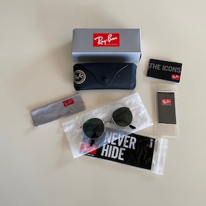 May include: A set of Ray-Ban sunglasses with a black frame and green lenses. The sunglasses are packaged in a black case with a Ray-Ban logo. The package also includes a gray microfiber cleaning cloth, a small black Ray-Ban card, and a white paper bag with the text "Never Hide" printed on it.