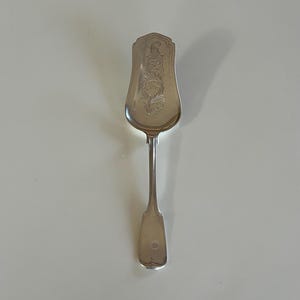 May include: A silver serving spoon with an ornate, floral design on the wide, rectangular head. The handle is slender and tapers to a rounded end. The spoon is likely vintage or antique.