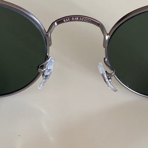 May include: Close-up of the inside of a pair of silver-toned round sunglasses with green lenses. The sunglasses have a Ray-Ban logo and the text "RAY-BAN 50□21" on the bridge.