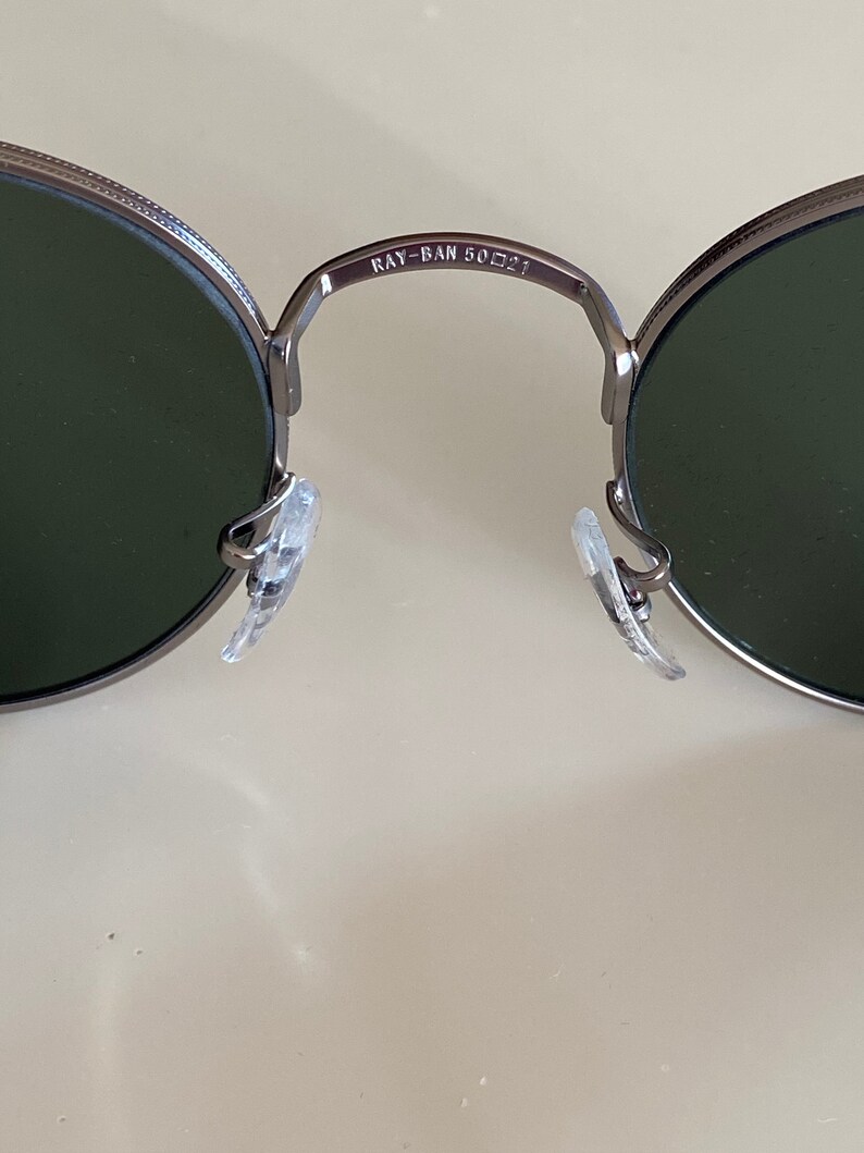 May include: Close-up of the inside of a pair of silver-toned round sunglasses with green lenses. The sunglasses have a Ray-Ban logo and the text "RAY-BAN 50□21" on the bridge.