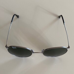 May include: A pair of round, dark gray sunglasses with silver frames and the Ray-Ban logo on the left lens.