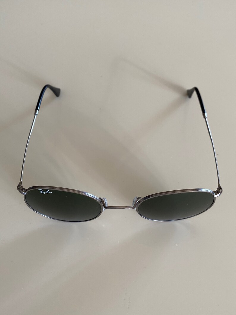 May include: A pair of round, dark gray sunglasses with silver frames and the Ray-Ban logo on the left lens.