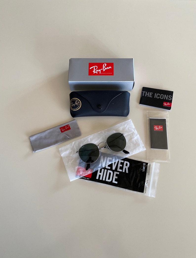 May include: A set of Ray-Ban sunglasses with a black frame and green lenses. The sunglasses are packaged in a black case with a Ray-Ban logo. The package also includes a gray microfiber cleaning cloth, a small black Ray-Ban card, and a white paper bag with the text "Never Hide" printed on it.