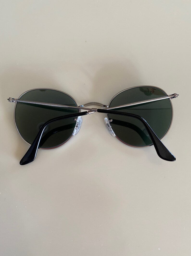 May include: A pair of round, silver-framed sunglasses with dark green lenses. The sunglasses have black arms.