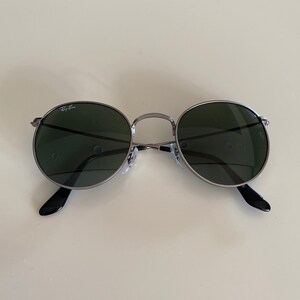 May include: A pair of round, dark green sunglasses with a silver frame and black arms. The sunglasses have the Ray-Ban logo on the left lens.