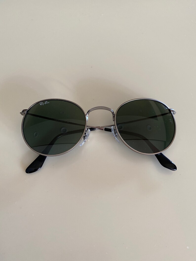 May include: A pair of round, dark green sunglasses with a silver frame and black arms. The sunglasses have the Ray-Ban logo on the left lens.