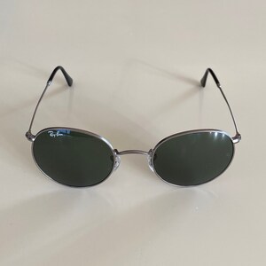 May include: Silver-framed round sunglasses with dark green lenses. The sunglasses have the Ray-Ban logo on the left lens.