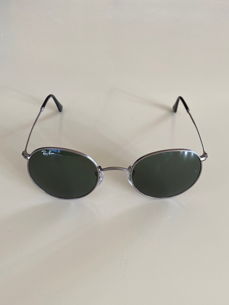 May include: Silver-framed round sunglasses with dark green lenses. The sunglasses have the Ray-Ban logo on the left lens.