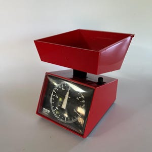 Puede incluir: Una báscula de cocina roja con un dial negro y una bandeja roja. El dial tiene marcas para libras y onzas.