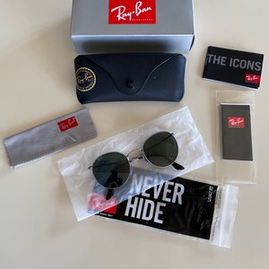 May include: A set of Ray-Ban sunglasses with green lenses and a silver frame. The sunglasses are packaged in a black case with a Ray-Ban logo. The set also includes a gray microfiber cleaning cloth, a black and white sticker that says "NEVER HIDE", and a small black and white card with the Ray-Ban logo.