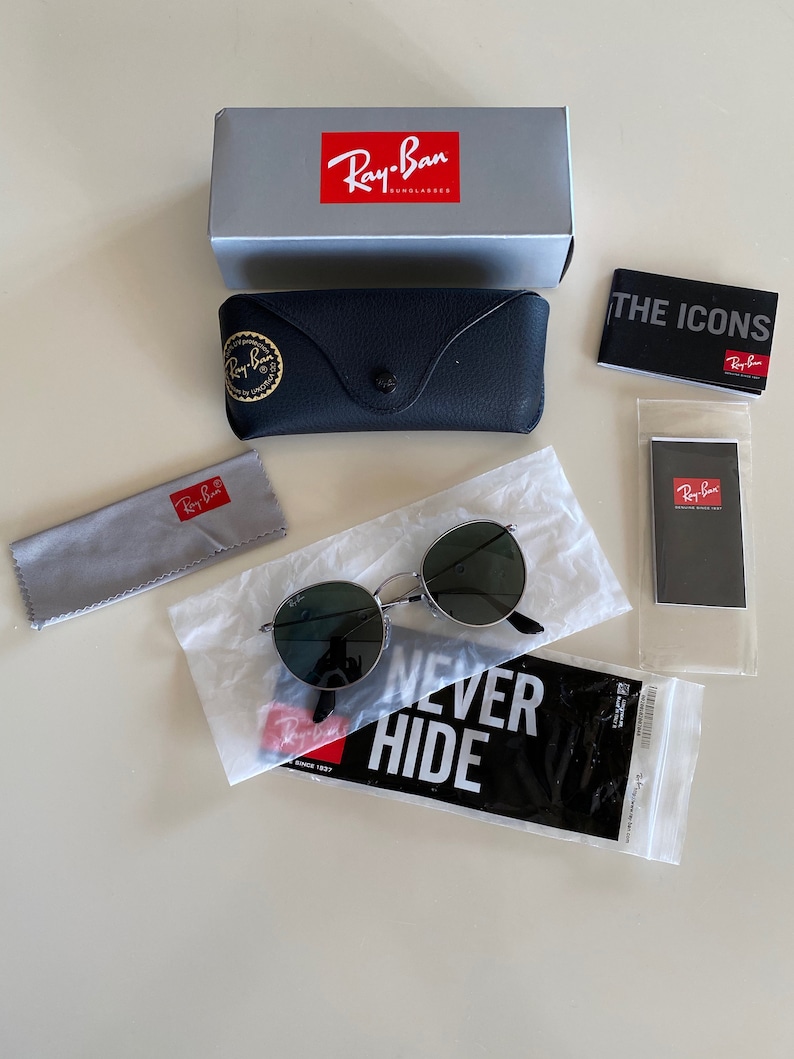 May include: A set of Ray-Ban sunglasses with green lenses and a silver frame. The sunglasses are packaged in a black case with a Ray-Ban logo. The set also includes a gray microfiber cleaning cloth, a black and white sticker that says "NEVER HIDE", and a small black and white card with the Ray-Ban logo.