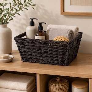 May include: A dark brown woven rectangular storage basket containing two folded beige towels and a round yellow sponge. Inside the basket are two pump bottles, one matte beige and one amber glass with a label reading meraki hand lotion.