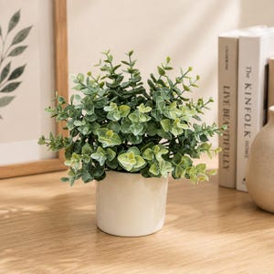 May include: A small artificial eucalyptus plant with rounded green leaves arranged in a dense, bushy cluster. The plant is housed in a simple, smooth, cylindrical white ceramic pot that sits upright on a flat surface.