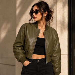 May include: An olive green cropped bomber jacket with a smooth finish, featuring a front zipper closure, ribbed cuffs, and a ribbed collar. The jacket is worn over a black crop top and black denim jeans, paired with dark-tinted cat-eye sunglasses.
