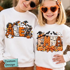 May include: Two white long-sleeve sweatshirts featuring custom Halloween-themed graphics. One sweatshirt displays the name ALEX in orange and black plaid letters with ghosts, bats, and pumpkins. The other displays EMMA with similar motifs.