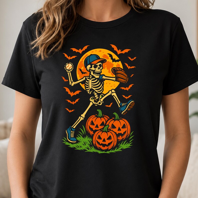 May include: A black t-shirt featuring a graphic of a skeleton wearing a baseball cap and glove, throwing a baseball. The skeleton is positioned above three jack-o'-lanterns on green grass, set against a large yellow moon and orange bats.