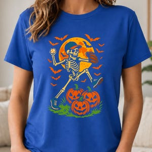 May include: A royal blue t-shirt featuring a graphic print of a skeleton wearing a baseball cap and glove, throwing a baseball. The design includes a large yellow full moon, orange bats, and three jack-o'-lanterns on a patch of green grass.