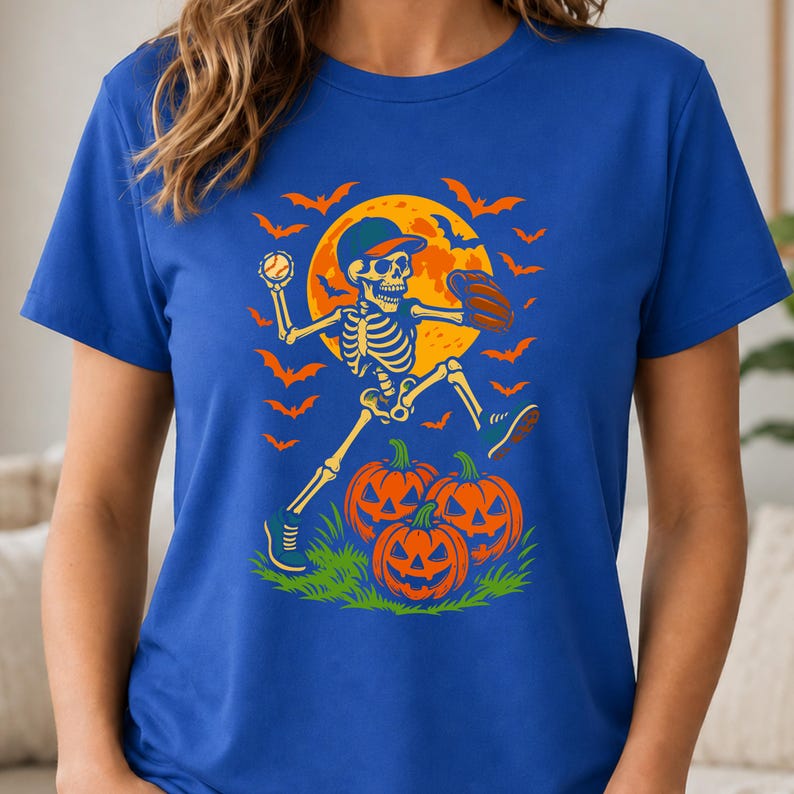 May include: A royal blue t-shirt featuring a graphic print of a skeleton wearing a baseball cap and glove, throwing a baseball. The design includes a large yellow full moon, orange bats, and three jack-o'-lanterns on a patch of green grass.