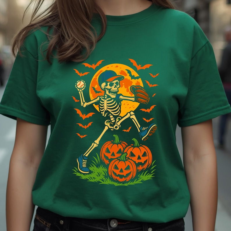 May include: A dark green short-sleeve t-shirt featuring a graphic of a skeleton wearing a baseball cap and glove, throwing a baseball. The design includes a large yellow moon, orange bats, and three jack-o'-lanterns on green grass.