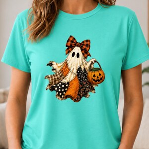 May include: A bright turquoise t-shirt featuring a central graphic of a ghost wearing a plaid bow. The ghost is composed of various orange, black, and white patterned fabric patches and is holding a small orange jack-o'-lantern pumpkin bucket.