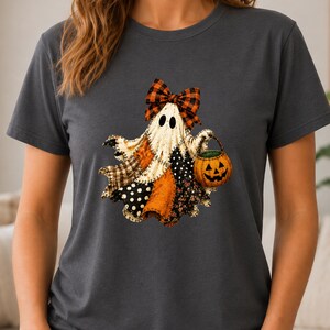 May include: A dark gray short-sleeve t-shirt featuring a graphic of a ghost wearing an orange and black plaid bow. The ghost has a patchwork body with various patterns and holds a small orange jack-o'-lantern bucket with a black face.