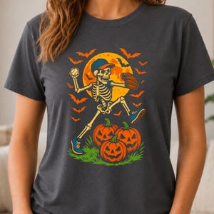 May include: A dark gray t-shirt featuring a graphic of a skeleton wearing a baseball cap and glove, throwing a baseball. The skeleton is positioned in front of a large yellow moon, surrounded by orange bats, with three jack-o'-lanterns below.