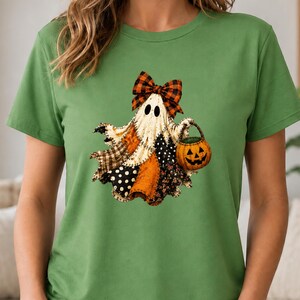 May include: A green short-sleeved t-shirt featuring a graphic of a ghost wearing a plaid bow. The ghost has a patchwork-style body with various patterns and is holding a small orange jack-o'-lantern bucket for trick-or-treating.