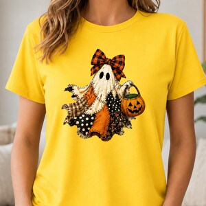 May include: A bright yellow short-sleeved t-shirt featuring a graphic of a ghost made of various patterned fabric patches. The ghost wears a large orange and black plaid bow on its head and carries a small orange jack-o'-lantern bucket.