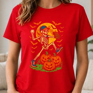 May include: A bright red t-shirt featuring a graphic print of a skeleton wearing a baseball cap and glove, throwing a baseball. The design includes a large yellow full moon, flying bats, and three orange jack-o'-lanterns on green grass.