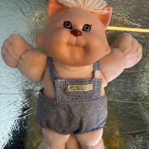 May include: A plush cat doll with a peach-coloured vinyl face, white yarn hair, and dark brown eyes and nose. The doll wears blue denim overalls with a small rectangular tag on the chest that features the text Cabbage Patch Kids.