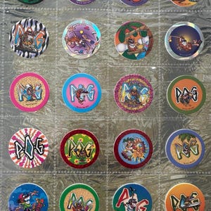 May include: A collection of twenty circular cardboard POG game discs arranged in a plastic storage sheet. Each disc features colorful cartoon illustrations, various graphic designs, and the POG logo, showcasing diverse artistic styles and themes.