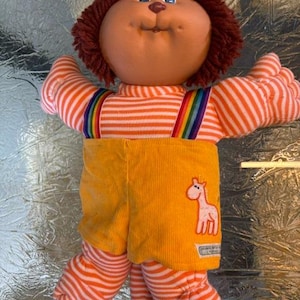 Peut inclure: Une poupée en peluche avec un visage rond bronzé, des yeux bleus et des cheveux en laine brune. La poupée porte un haut à manches longues rayé orange et blanc avec des bretelles arc-en-ciel et une salopette en velours côtelé jaune.