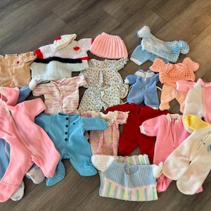 May include: A collection of assorted doll clothing including various knitted sweaters, fleece footed pyjamas in pink, blue, red, and white, a pink skirt, and patterned dresses. The garments feature diverse textures, colours, and closure styles.