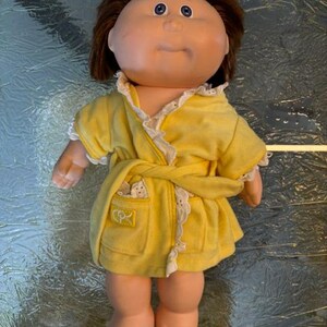 May include: A doll with brown hair and blue eyes wearing a yellow robe with white lace trim. The robe features a tie waist and a small pocket on the front with the letters CPK embroidered in yellow thread on a square patch.