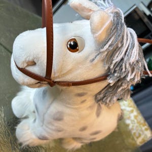 Peut inclure: Une peluche en forme de cheval blanc avec des taches grises, dotée d'une bride en similicuir marron et d'une crinière en laine grise et blanche. Le cheval a de grands yeux marron et une fourrure blanche douce avec une bride autour de la tête.