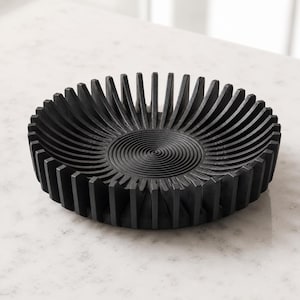 May include: A circular black bowl featuring a series of vertical, radiating fins along the outer edge and a concentric, spiraling circular pattern etched into the flat base of the interior, creating a textured and geometric appearance.