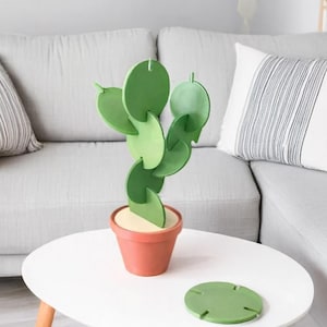 May include: A green cactus-shaped sculpture constructed from interlocking circular discs, seated in a terracotta-colored pot with a light-colored base. An additional detached green circular disc lies flat on the surface next to the potted sculpture.