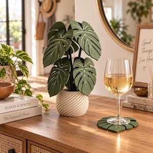 Könnte beinhalten: Eine dekorative künstliche Monstera-Pflanze mit dunkelgrünen Blättern und weißen Blattadern in einem cremefarbenen, spiralförmig gerillten zylindrischen Topf. Ein passender blattförmiger Untersetzer steht unter einem Glas Weißwein.