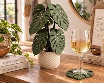 May include: A decorative artificial monstera plant with dark green leaves featuring white vein details, potted in a cream-coloured, spiralled, cylindrical base. A matching dark green leaf-shaped coaster sits under a clear glass of white wine.