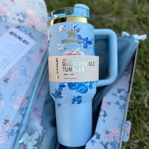 May include: A light blue Stanley Quencher H2.0 tumbler with a 40 oz capacity, featuring a floral pattern in shades of blue and white. The tumbler has a gold-tone rim, a matching handle, and a paper band labeled The Quencher H2.0 Tumbler 40 oz.