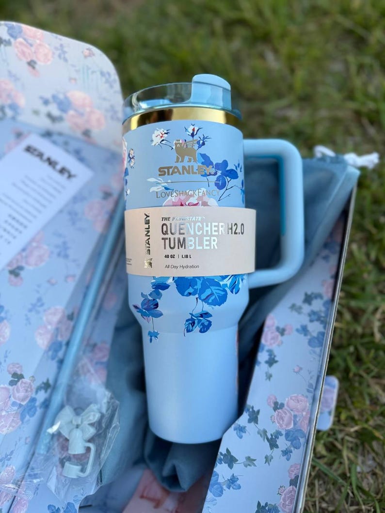 May include: A light blue Stanley Quencher H2.0 tumbler with a 40 oz capacity, featuring a floral pattern in shades of blue and white. The tumbler has a gold-tone rim, a matching handle, and a paper band labeled The Quencher H2.0 Tumbler 40 oz.
