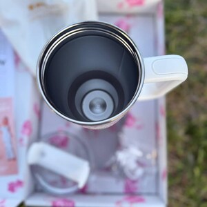 Op de afbeelding: Een witte roestvrijstalen beker met handvat, van bovenaf gezien. De binnenkant is donkergrijs met een metalen rand. Een doorzichtig plastic deksel met een witte schuifsluiting ligt ernaast in een witte doos met roze bloemmotieven.