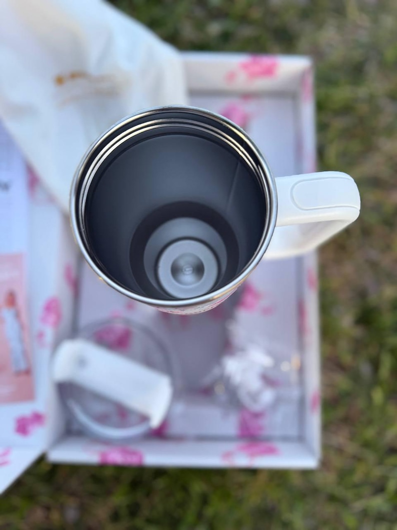 Op de afbeelding: Een witte roestvrijstalen beker met handvat, van bovenaf gezien. De binnenkant is donkergrijs met een metalen rand. Een doorzichtig plastic deksel met een witte schuifsluiting ligt ernaast in een witte doos met roze bloemmotieven.