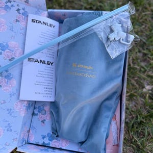 May include: A blue Stanley and LoveShackFancy branded fabric pouch inside a floral patterned box. The set includes a light blue straw and a small white plastic accessory in a clear bag, alongside a Stanley instruction manual with printed text.