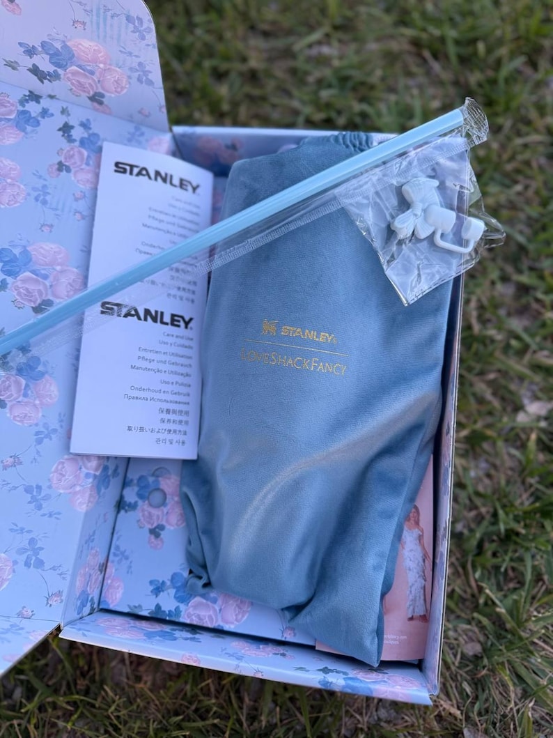 May include: A blue Stanley and LoveShackFancy branded fabric pouch inside a floral patterned box. The set includes a light blue straw and a small white plastic accessory in a clear bag, alongside a Stanley instruction manual with printed text.