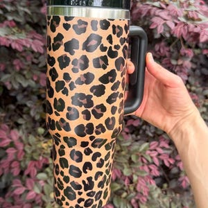 May include: A tall, cylindrical stainless steel tumbler featuring a tan and black leopard print pattern. The drinkware includes a black plastic lid and a matching black side handle, with a tapered base design for a secure grip and easy storage.
